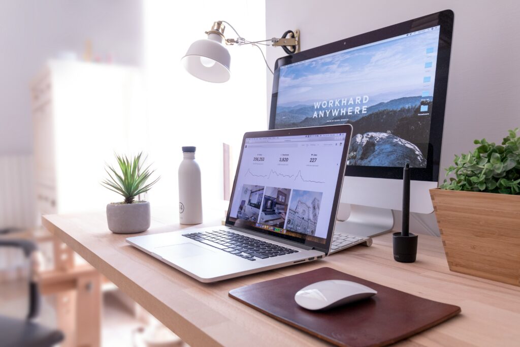 A tidy desk with a laptop, desktop monitor displaying WORK HARD ANYWHERE, a desk lamp, mouse, mouse pad, potted plants, a water bottle, and a stylus holder, all arranged neatly in a well-lit room.