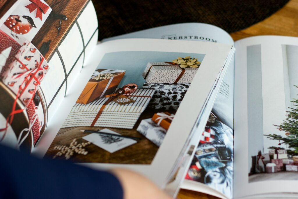 An open magazine displays pages with photos of wrapped Christmas presents and a decorated Christmas tree. A persons hand is visible at the bottom of the image, holding the magazine.