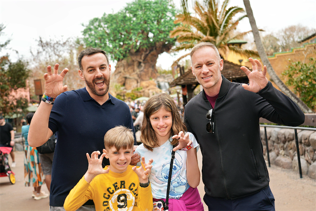 Four people, two adults and two children, pose together outdoors, smiling and making playful claw gestures with their hands. Trees and theme park scenery are visible in the background.