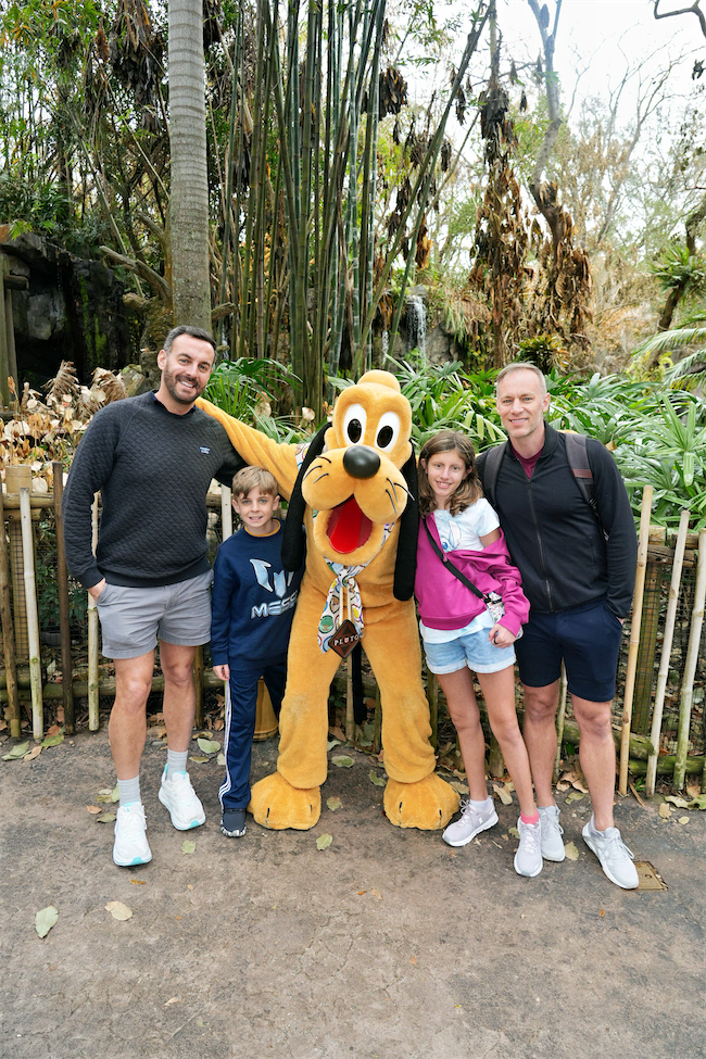 Two adults and two children pose and smile with a person in a Pluto costume at an outdoor theme park, surrounded by lush greenery and tropical plants.