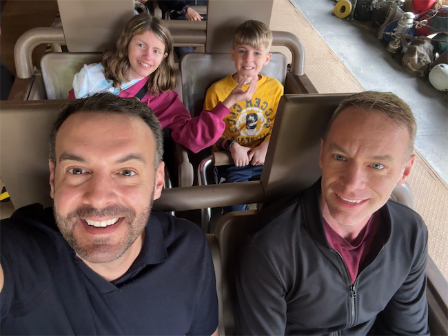 Two adults and two children sit together on a theme park ride, smiling at the camera. The adults are in front and the children are behind them, all appearing happy and excited for the ride.
