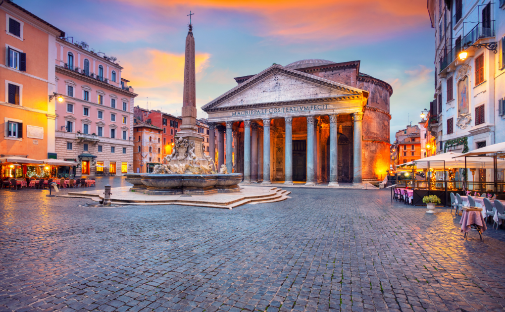 A view of the Pantheon in Rome at sunset, with the ancient columns and fountain in front, surrounded by outdoor café seating and colorful buildings under a colorful sky.