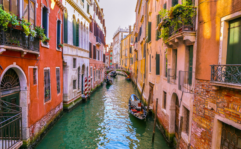 A narrow canal in Venice, Italy, lined with colorful historic buildings, balconies with flowers, and boats including a gondola gliding through the water under a small bridge.