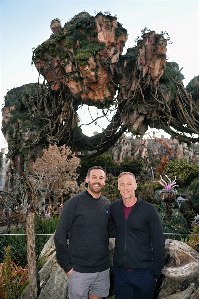 Two people stand smiling in front of a lush, fantastical landscape with floating mountains and vibrant plants, resembling the world of Pandora from the movie Avatar.