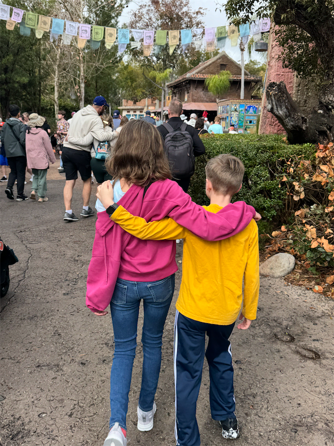 Two children, one in a pink jacket and the other in a yellow shirt, walk arm in arm down a path in a busy outdoor area with people, trees, and colorful flags overhead.