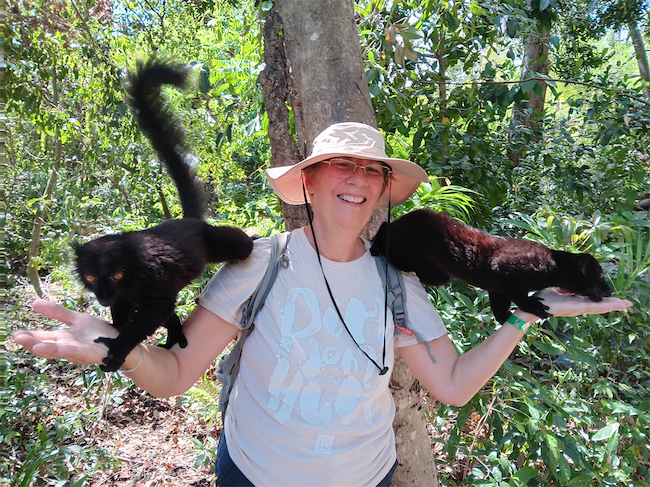 A smiling person wearing a hat stands in a lush, green forest with a black lemur on each arm, showing that not all points are equal—one lemur climbs their left arm while the other perches playfully on their right hand.