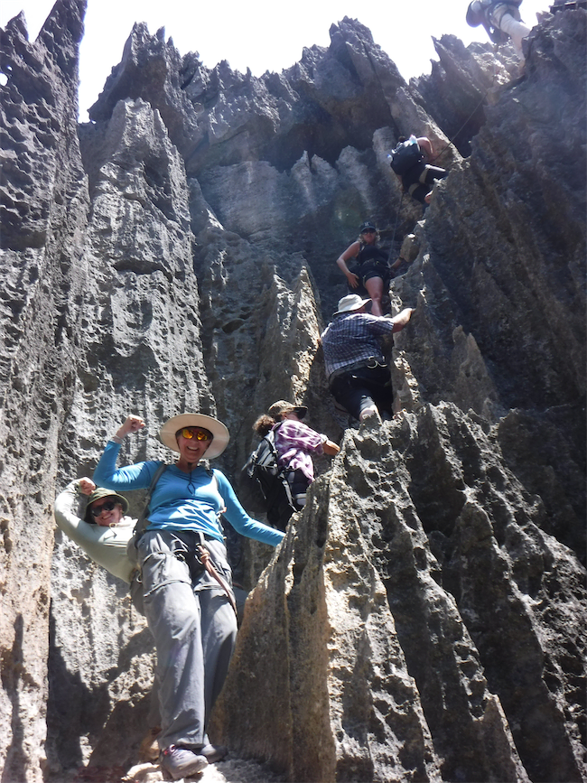 A group of people climbs steep, jagged limestone rock formations, with some climbers smiling and posing for the camera while others ascend carefully in the background, showing that not all points are equal on this challenging route under a bright sky.