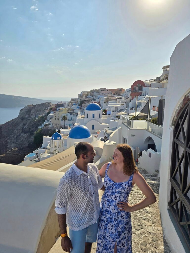 A couple stands close together on a sunny stone path, with the iconic white buildings and blue domes of Santorini, Greece, in the background. The sky is clear, and the sea is visible along the cliffside.