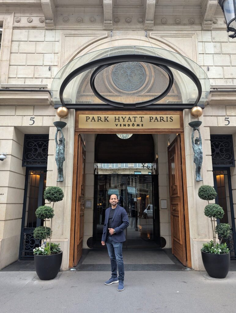A person stands smiling in front of the entrance to Park Hyatt Paris-Vendôme, flanked by potted plants and ornate details on the building's facade.