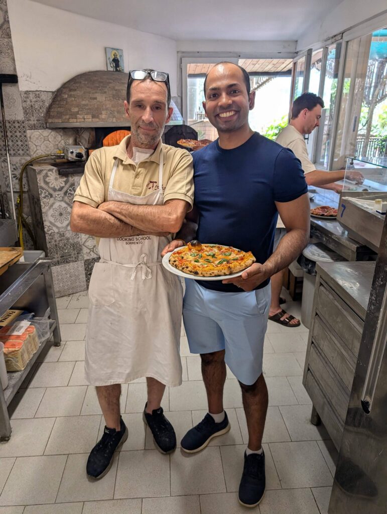 Two men stand in a pizzeria kitchen; one wears an apron and folds his arms, while the other smiles and holds a fresh pizza. Other workers prepare food in the background.