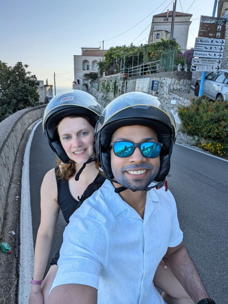 Two people wearing helmets, smiling while riding a scooter on a scenic road lined with buildings and greenery, with street signs visible in the background.