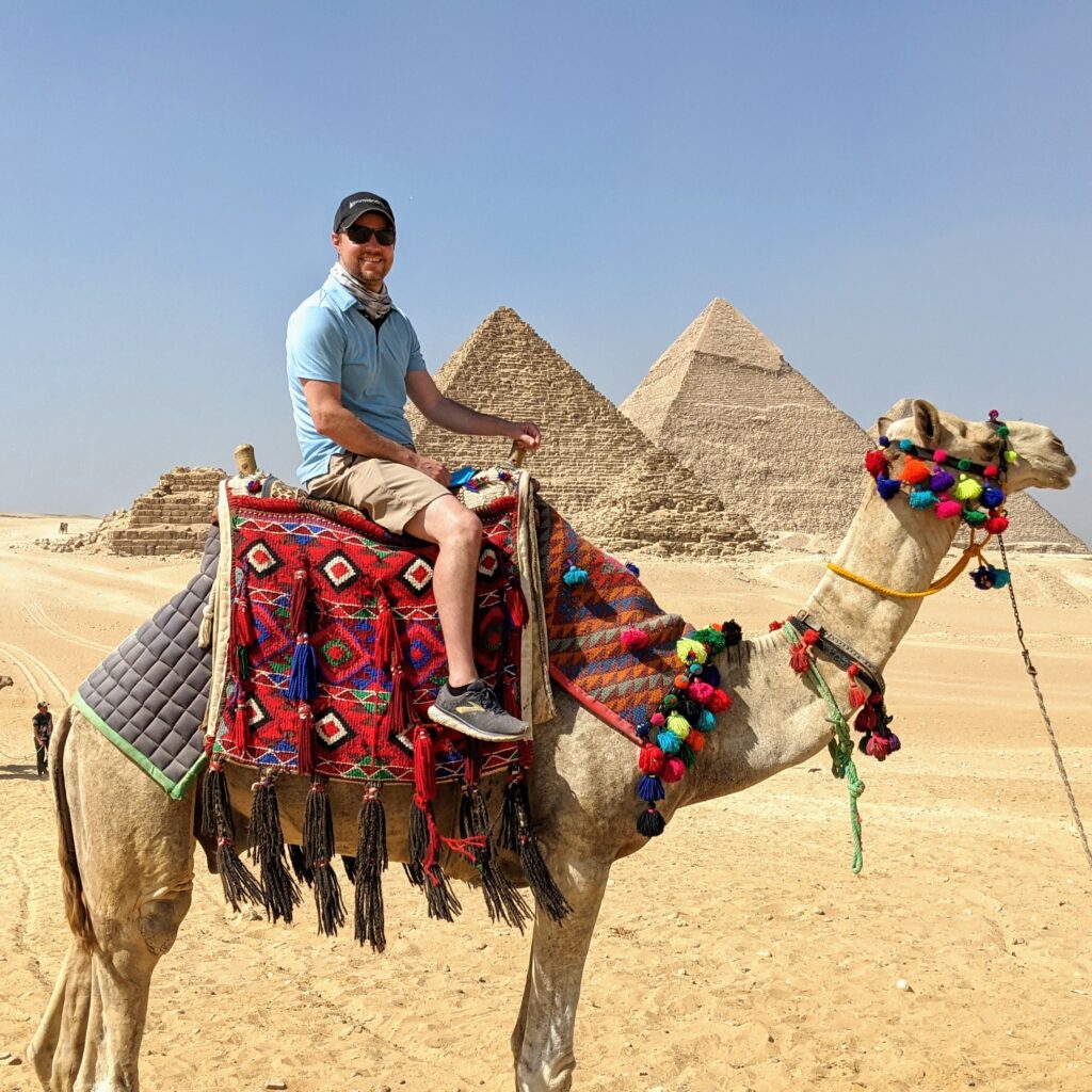 A man in sunglasses and a blue shirt rides a decorated camel near the Pyramids of Giza under a clear sky. The desert and pyramids are visible in the background.