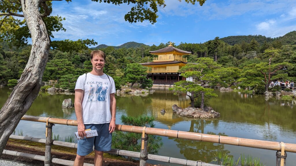 A man in a Coldplay T-shirt stands by a bamboo fence in front of the Golden Pavilion (Kinkaku-ji) in Kyoto, Japan, surrounded by trees and a reflective pond under a blue sky.