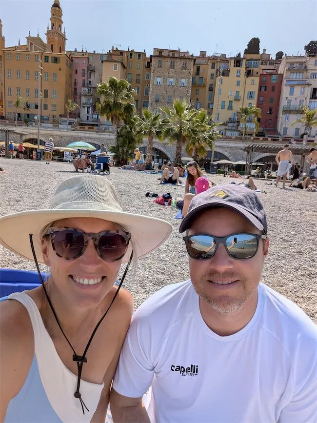 Smiling couple wearing sunglasses and hats pose for a selfie on a pebbly beach, with colorful buildings, palm trees, and other beachgoers in the background.