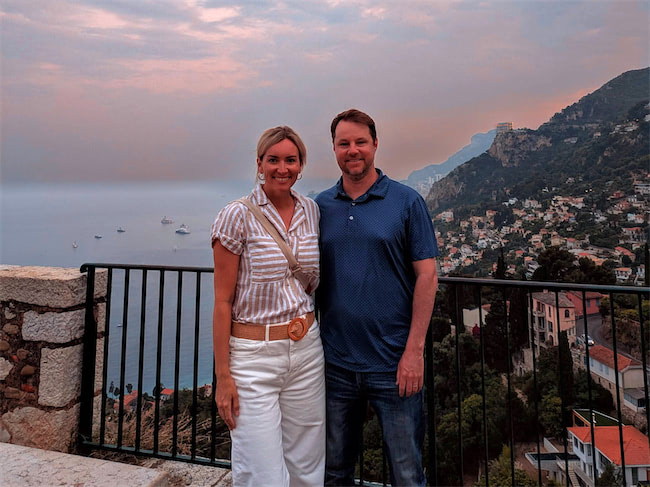 A woman and man stand smiling on a balcony overlooking a coastal town and sea at sunset, with hills and boats visible in the background.