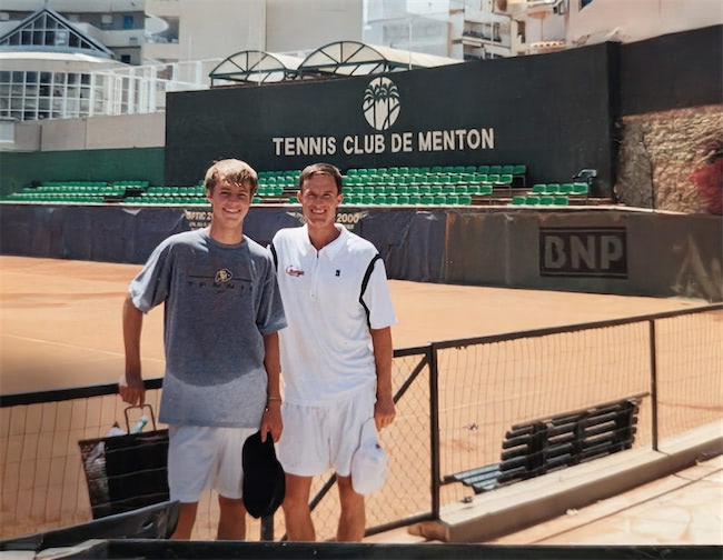 Two men in sportswear stand on a clay tennis court in front of green stands and a sign reading Tennis Club de Menton. Both are smiling and holding tennis equipment on a sunny day.