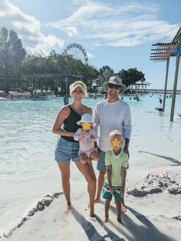 A family of four poses on a sandy beach by a pool, with trees and a Ferris wheel in the background. Two adults stand behind a young child and toddler. All are wearing sun hats and sunglasses, enjoying a sunny day.
