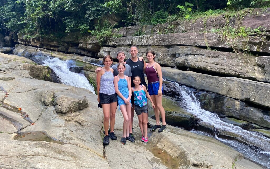 Six people stand together on rocks beside a small waterfall in a lush, forested area. They are all wearing casual, sporty clothing and smiling at the camera, enjoying an adventure where not all points are equal—some experiences simply stand out above the rest.