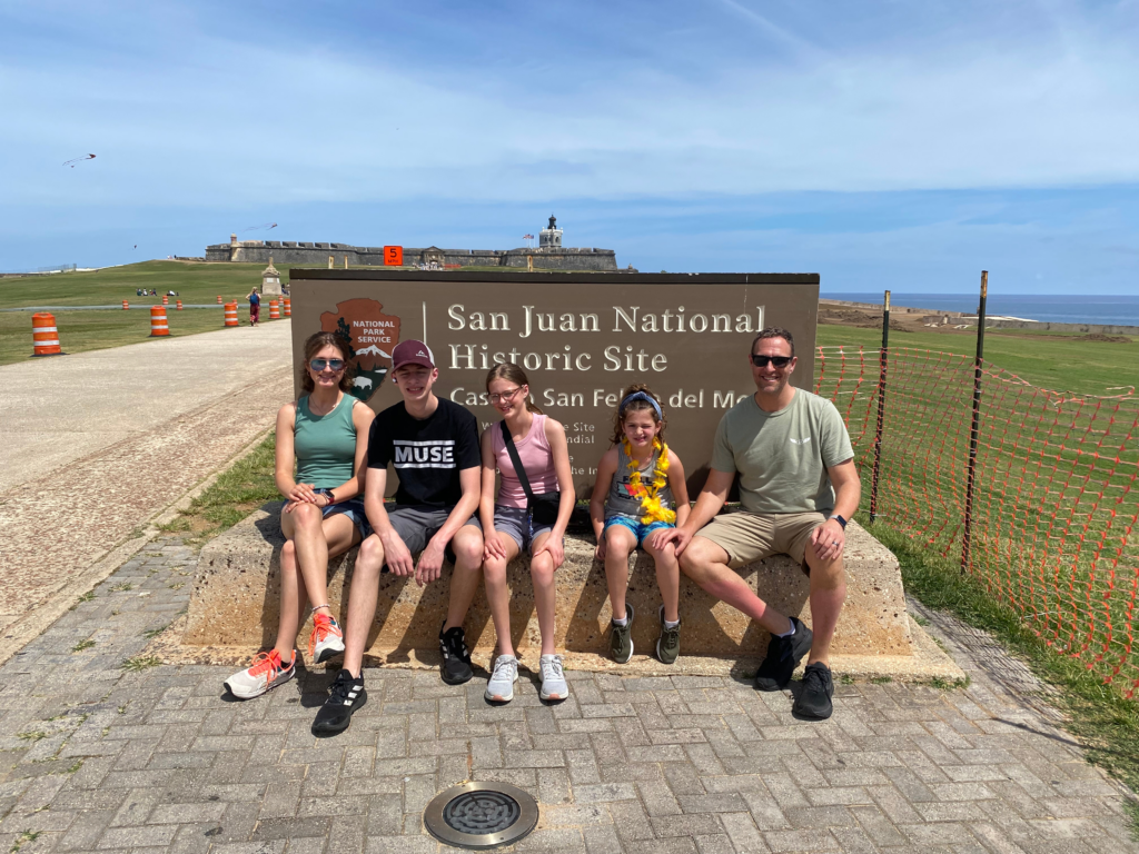 Five people sit on a concrete block in front of a San Juan National Historic Site sign, with a green lawn, historic fort, and blue sky in the background—a scene that subtly reminds us that not all points are equal when it comes to choosing the perfect spot to take in this breathtaking view.