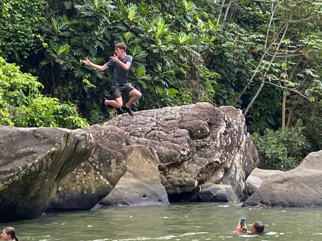 A person mid-air jumps off a large rock into a river surrounded by lush greenery, perfectly capturing the exhilaration of adventure—while another person in the water takes a photo with a smartphone, reminding us that not all points are equal when it comes to experiencing life’s unforgettable moments.